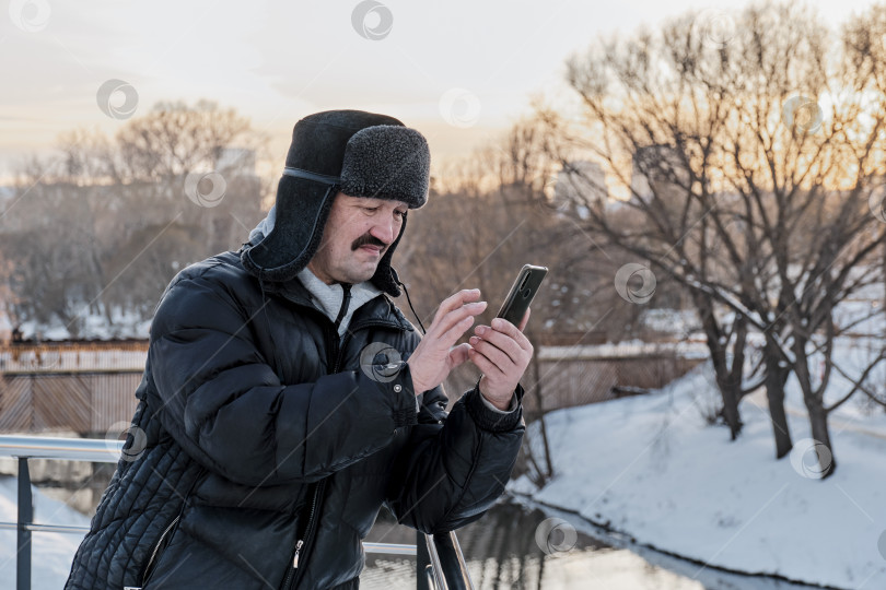 Скачать Пожилой азиат в зимней шапке-ушанке пользуется мобильным телефоном в парке, фотографируя природу фотосток Ozero