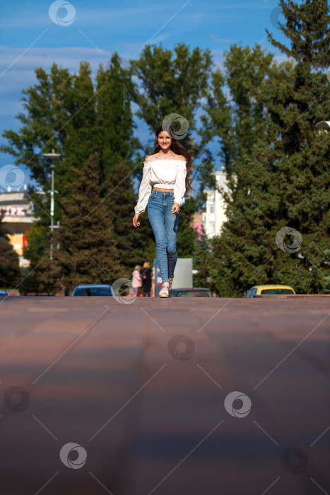 Скачать Молодая красивая брюнетка в джинсах и белой блузке прогуливается по летней улице фотосток Ozero