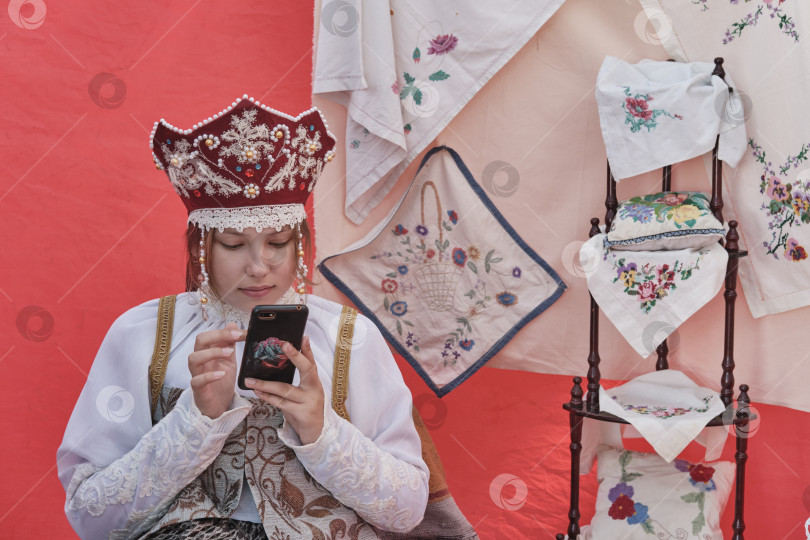 Скачать Девушка в стилизованной русской традиционной одежде прокручивает экран мобильного телефона на сельском празднике. фотосток Ozero