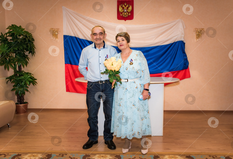 Скачать Пожилая российская пара в возрасте 75 лет, празднующая золотую свадьбу или 50-летнюю годовщину свадьбы в реестре регистрации браков, на их медалях на русском языке написано "50-летняя годовщина свадьбы". фотосток Ozero