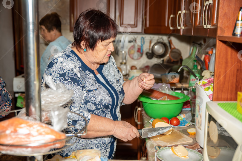 Скачать 70-летняя женщина нарезает колбасу на деревянной разделочной доске на кухне, пробуя кусочек. Ее подруга помогает готовить праздничный стол. Винтажная кухня и подвесные сковородки фотосток Ozero