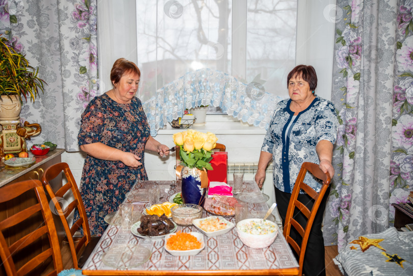 Скачать Две русские женщины, 70 и 60 лет, готовят праздничный стол с традиционными блюдами, такими как салаты, котлеты и мясо. Старинная деревенская кухня и уютная атмосфера создают настроение для веселого праздничного застолья фотосток Ozero