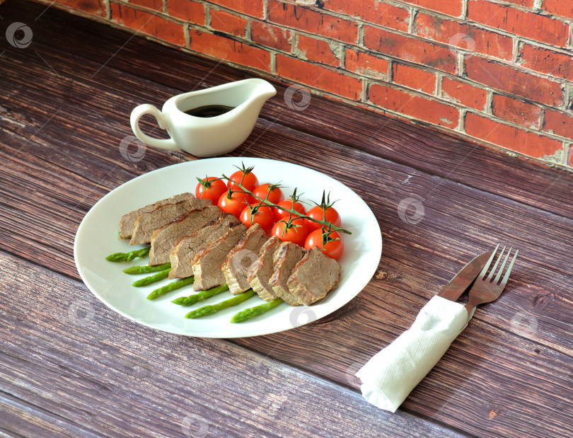 Скачать Приготовленный на гриле говяжий стейк, нарезанный кусочками, с помидорами черри и спаржей на деревянном столе, рядом с чашкой с соусом и столовыми приборами. фотосток Ozero