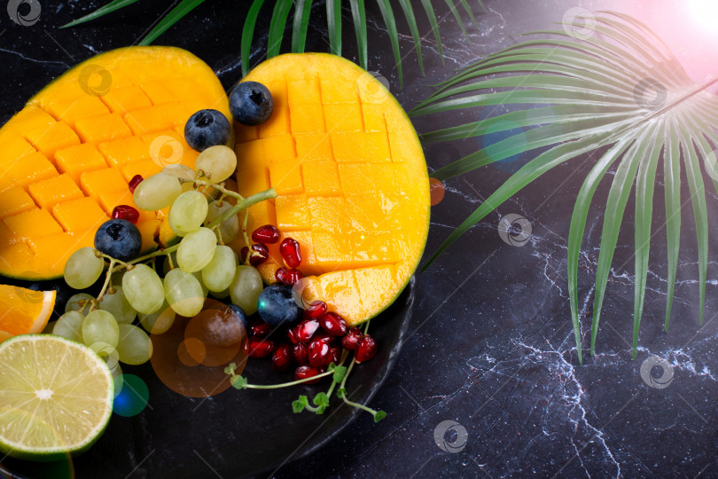 Скачать Ассорти из тропических фруктов подается на темном блюде: манго, виноград, лайм, черника и гранатовые косточки на черном столе. фотосток Ozero