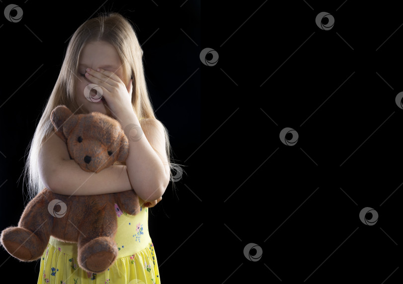 Скачать Маленькая девочка обнимает игрушечного мишку на темном фоне, плачет и закрывает лицо руками фотосток Ozero