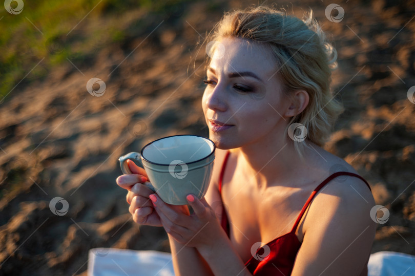 Скачать Красивая девушка, укрытая пледом вечером на закате, держит кружку чая двумя руками на фоне моря. фотосток Ozero