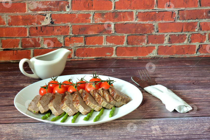 Скачать Запеченная говядина, нарезанная кусочками, с помидорами черри и отварной спаржей на деревянном столе, рядом с соусником и столовыми приборами в салфетке. фотосток Ozero