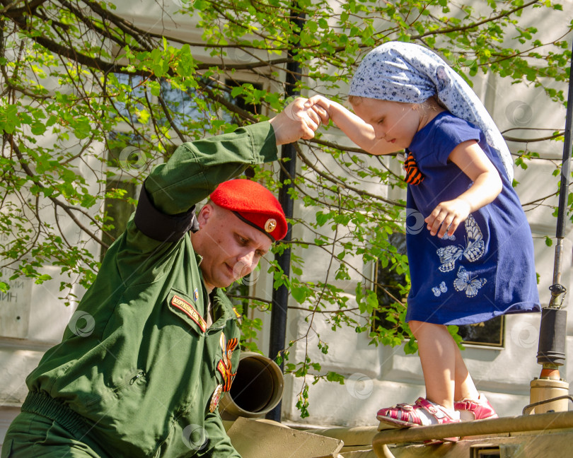 Скачать Ульяновск, Россия - 09 мая 2019 года: Выставка военной техники на центральной площади города. Солдат российской армии помогает маленькой девочке спуститься с боевой машины. фотосток Ozero