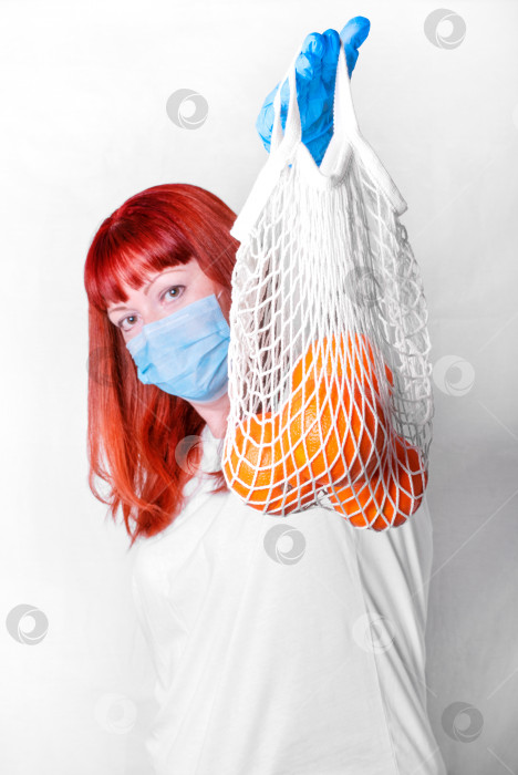 Скачать Рыжеволосая женщина в медицинской маске и перчатках держит апельсины в сетчатом пакете. Ходила за продуктами во время вспышки пандемии. фотосток Ozero
