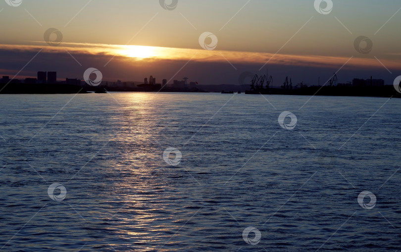 Скачать Закат на воде возле портового города. Темные силуэты кранов и домов вдали. Отражение солнца на волнах фотосток Ozero