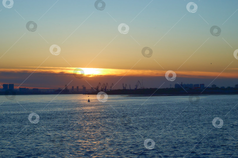 Скачать Закат на воде возле портового города. Темные силуэты кранов и домов вдали. Отражение солнца на волнах фотосток Ozero