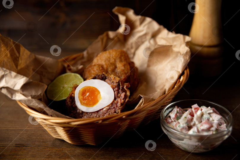 Скачать Порция шотландского яйца и соуса пико де галло подается на крафтовой бумаге и в плетеной корзинке. Жареная еда из паба, снятая в интерьере бара на фоне темного дерева. фотосток Ozero