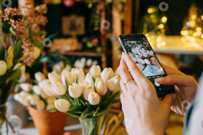 Скачать Крупный план человека, фотографирующего букет белых тюльпанов с помощью смартфона в магазине цветочных композиций. Фон с красивым освещением и цветочным декором создает теплую и располагающую атмосферу. фотосток Ozero