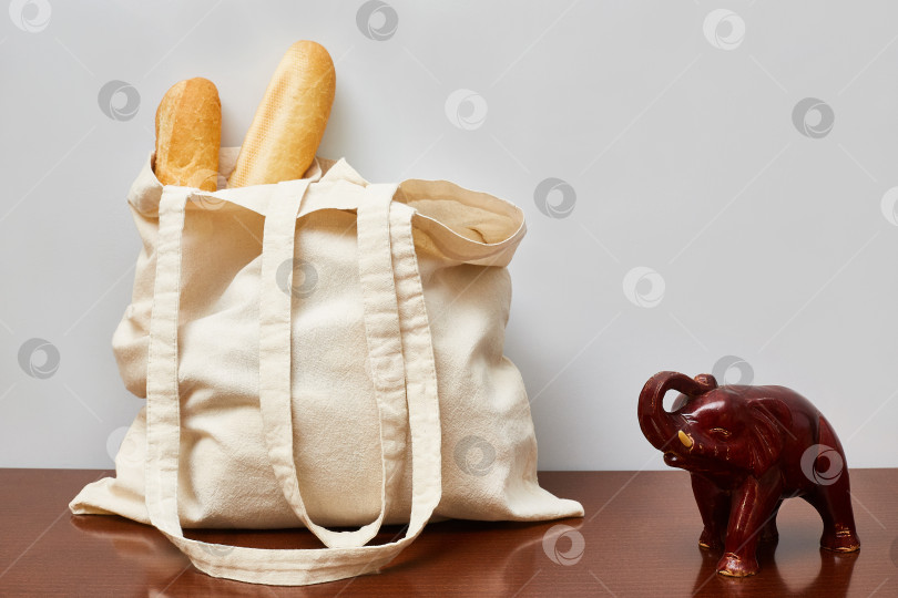 Скачать На комоде лежит сумка из эко-хлопка с батонами французского хлеба. Рядом стоит фигурка деревянного слоника. фотосток Ozero