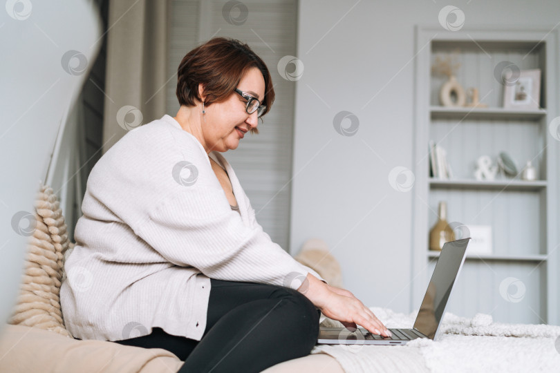 Скачать Улыбающаяся женщина средних лет плюс размер, работающая на ноутбуке дома фотосток Ozero