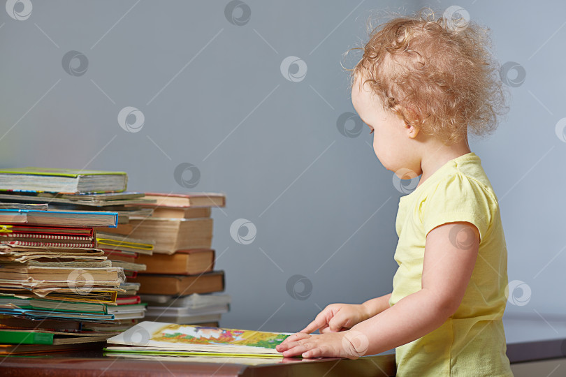 Скачать Ребенок смотрит на книгу. Маленький мальчик или девочка читает книгу и указывает на нее пальцем. Рядом с книгой большая стопка книг. Место для тетрадей. фотосток Ozero