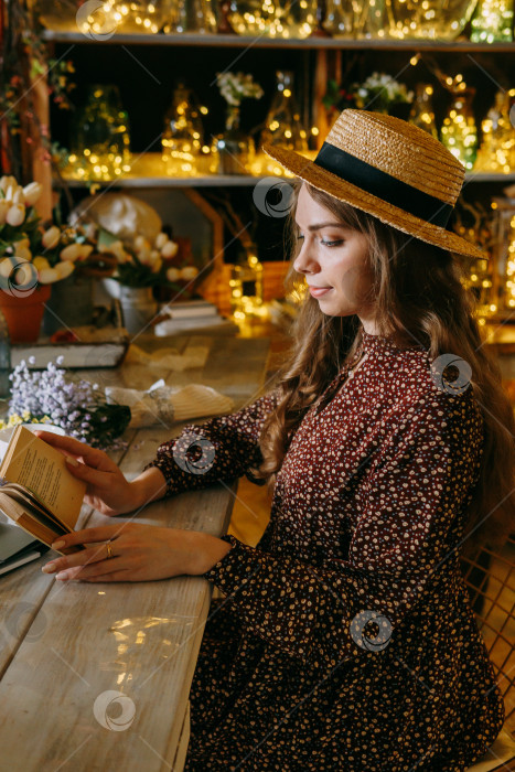 Скачать Женщина в соломенной шляпе и платье в цветочек читает книгу в теплой, красиво освещенной обстановке с цветами и очаровательным декором. фотосток Ozero