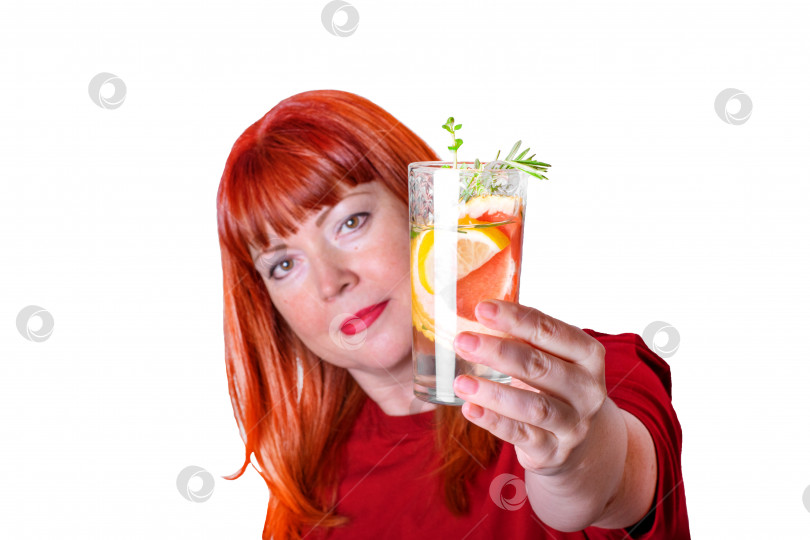 Скачать Бокал освежающего напитка с ломтиками лимона и грейпфрута предлагает рыжеволосая женщина средних лет в красной футболке, выделяющейся на белом фоне. фотосток Ozero