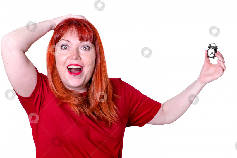 Скачать Рыжеволосая женщина средних лет в красной футболке держит в руках маленький будильник и кричит от шока. Концепция нехватки времени и опоздания. фотосток Ozero