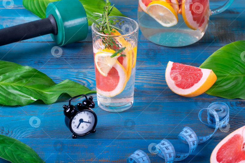Скачать Настоянная вода с грейпфрутом, лимоном и мятой в высоком стакане, будильник, зеленая гантель и измерительная лента на синем деревянном столе. фотосток Ozero