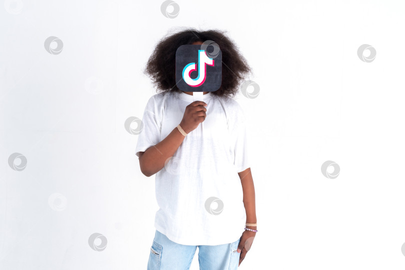 Скачать Подросток-афроамериканец держит в руках картинку с логотипом Tiktok на белом фоне. Подростки увлекаются новыми технологическими тенденциями - концепцией молодежи, технологий, общения и дружбы. Турция, Стамбул, 29 мая 2022 г. фотосток Ozero