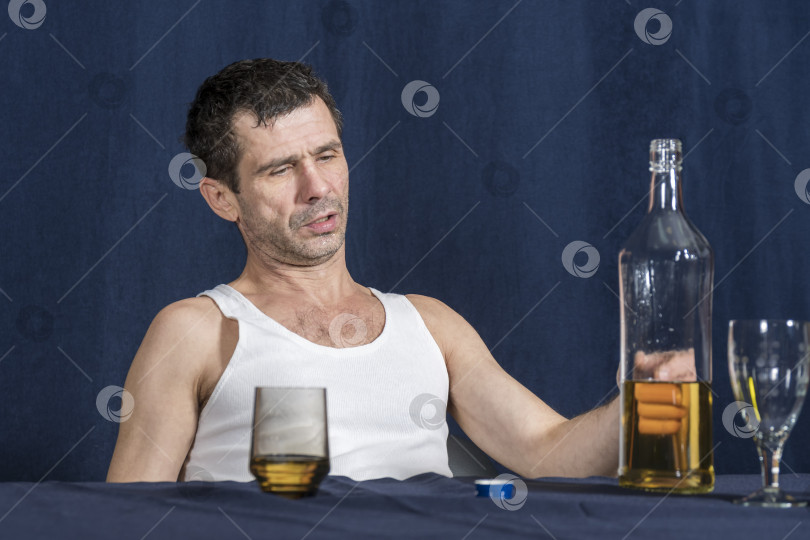 Скачать Пьяный, одинокий, но гордый мужчина сидит за столом и смотрит на бутылку алкоголя, стоящую перед ним. фотосток Ozero