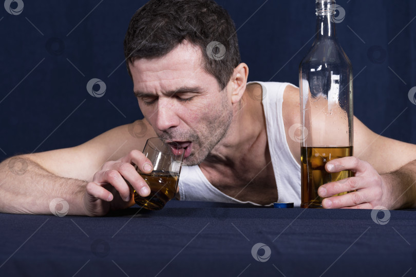 Скачать Пьяный одинокий мужчина лежит грудью на столе и пытается дотянуться ртом до стакана с алкоголем. Держит бутылку алкоголя в одной руке. фотосток Ozero