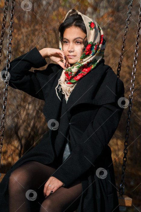 Скачать Красивая девушка сидит в парке на качелях, на голове у нее традиционный русский платок фотосток Ozero