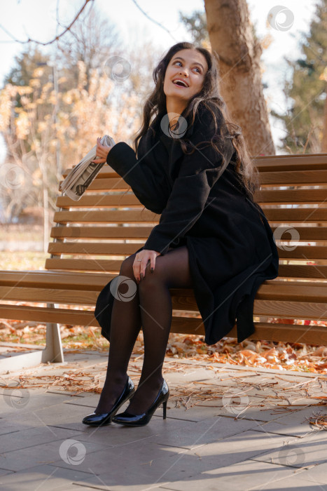 Скачать Девушка в черном пальто в парке сидит на скамейке и весело что-то говорит, размахивая газетой фотосток Ozero