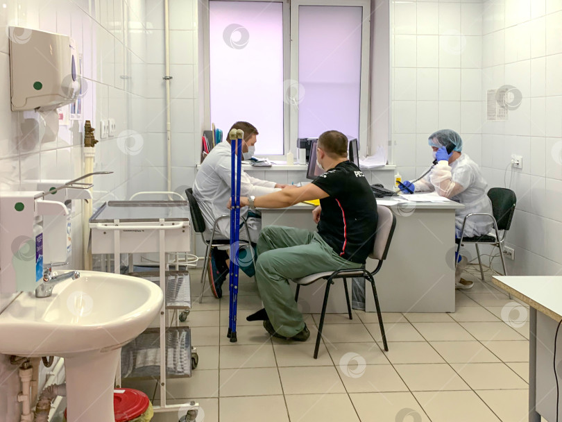 Скачать Мужчина со сломанной ногой и на костылях на приеме у врача во время пандемии коронавируса. Кабинет врача в больнице. Россия, Татарстан, 05 апреля 2020 г. фотосток Ozero