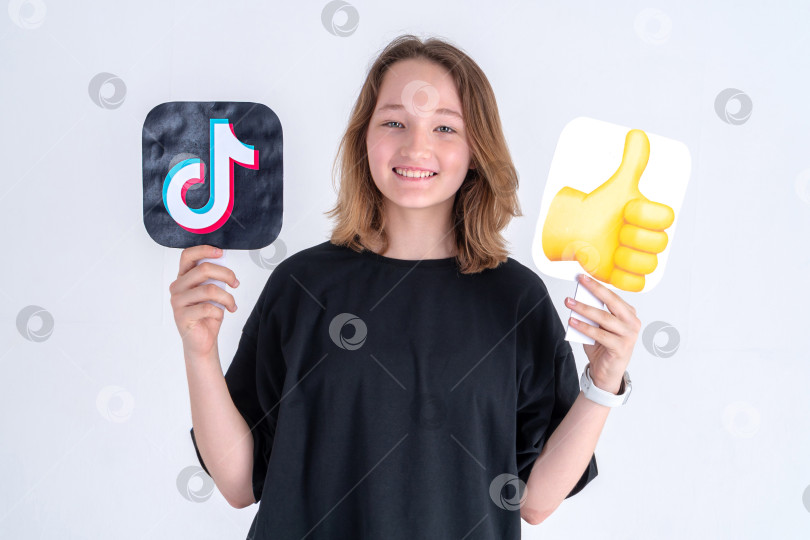 Скачать счастливая белая девочка-подросток в черной футболке с логотипом tiktok и эмодзи "большой палец вверх", выделенными на белом фоне. Турция, Стамбул, 29 мая 2022 г. фотосток Ozero