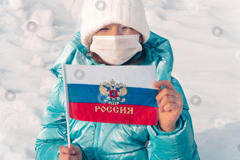 Скачать Девочка-подросток в маске с российским флагом в руках на фоне белого снега. Скажите "нет" короновирусу. Эпидемия гриппа и сезон заболеваний фотосток Ozero