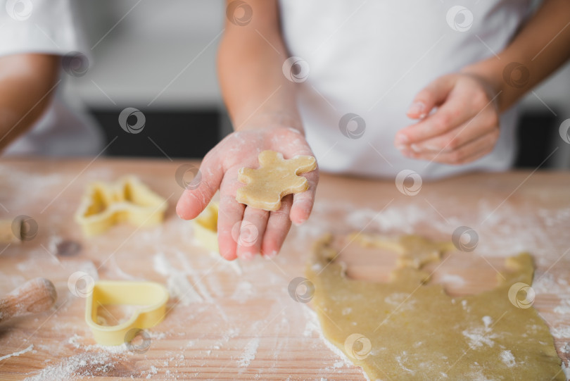 Скачать Крупный план детской ладошки с кусочком печенья из сырого теста, напоминающим медвежонка, на заднем плане кухонный стол, перепачканный мукой. фотосток Ozero