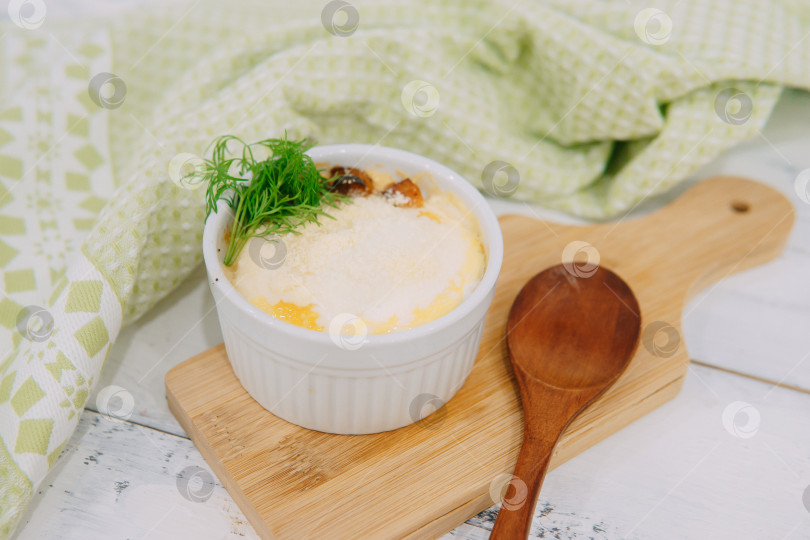 Скачать Вкусное запеченное блюдо, которое подается в белой формочке, украшенное свежим укропом, выкладывается деревянной ложкой на деревянную разделочную доску, создавая уютную кулинарную атмосферу в деревенском стиле. фотосток Ozero
