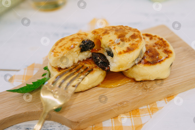 Скачать Творожные блинчики с изюмом и медом, наколотые золотой вилкой, подаются на деревянной доске на завтрак. Фотография закусочной фотосток Ozero