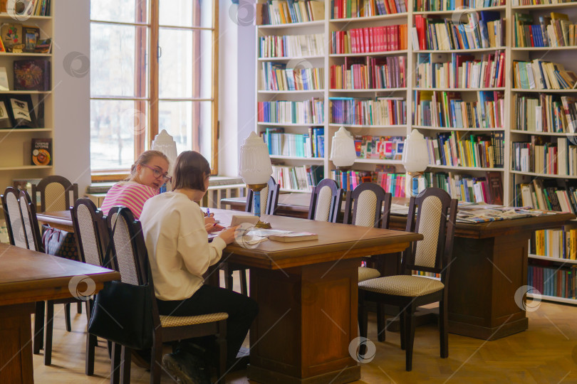 Скачать Молодые студентки сидят за столом в библиотеке и читают книгу фотосток Ozero