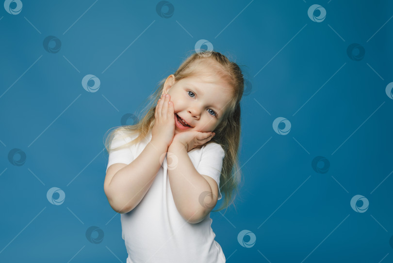 Скачать Веселая девочка 2-3 лет держит руки у лица и счастливо улыбается на голубом фоне. Счастливый ребенок радуется успехам и победам. фотосток Ozero
