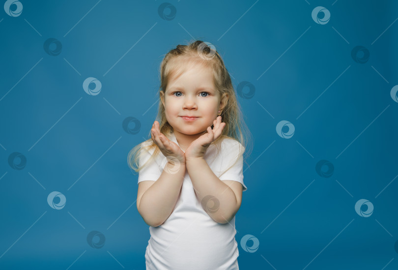 Скачать Веселая девочка 2-3 лет держит руки у лица и счастливо улыбается на голубом фоне. Счастливый ребенок радуется успехам и победам. фотосток Ozero
