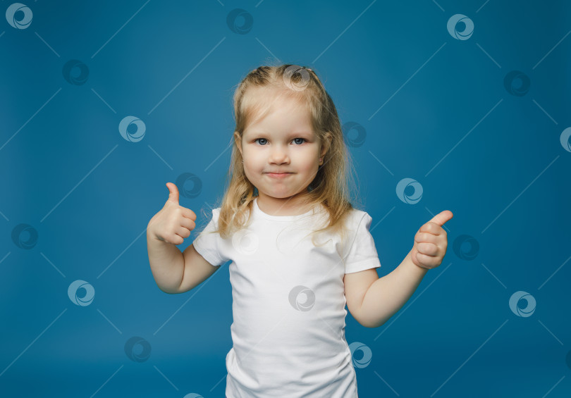 Скачать Фотография жизнерадостной счастливой девочки 2-3 лет, показывающей большой палец вверх, что означает "рекомендуется", выделенная на синем фоне. фотосток Ozero