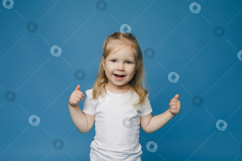 Скачать Фотография жизнерадостной счастливой девочки 2-3 лет, показывающей большой палец вверх, что означает "рекомендуется", выделенная на синем фоне. фотосток Ozero