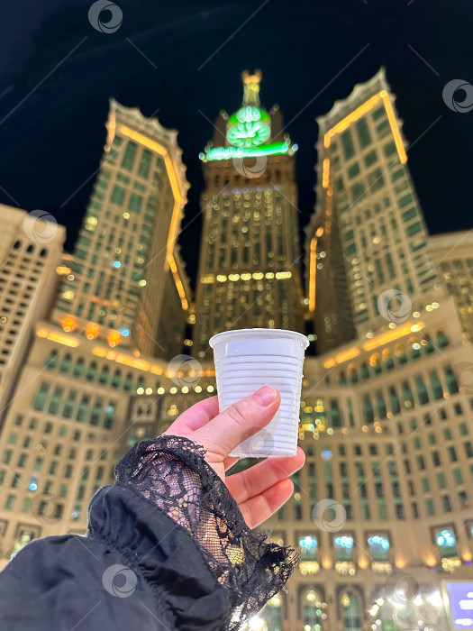 Скачать Исламский фон. Женская рука держит стакан со святой водой "замзам" на фоне знаменитой королевской башни с часами в Мекке. Место для текста. Абрадж Аль-Байт. Саудовская Аравия, Мекка, 24 ноября 2024 года, фотосток Ozero
