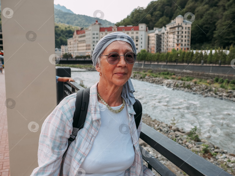 Скачать Портрет красивой пожилой женщины на отдыхе в горах у реки. 60-летняя женщина в очках, тюрбане и клетчатой рубашке. фотосток Ozero