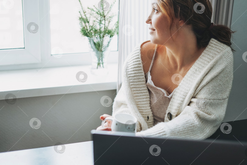 Скачать Привлекательная дружелюбная улыбающаяся брюнетка среднего возраста, пользующаяся ноутбуком дома, создает уютную атмосферу зимнего сезона фотосток Ozero