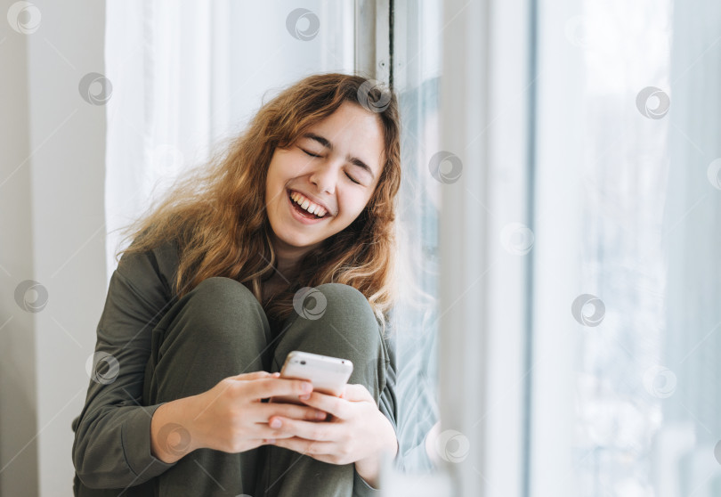 Скачать Красивая улыбающаяся девушка-подросток с вьющимися волосами, пользующаяся мобильным телефоном, сидя на подоконнике, удаленная связь фотосток Ozero