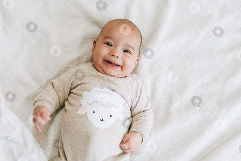 Скачать Милый улыбающийся мальчик 4-6 месяцев в бежевом комбинезоне на кровати естественных тонов фотосток Ozero