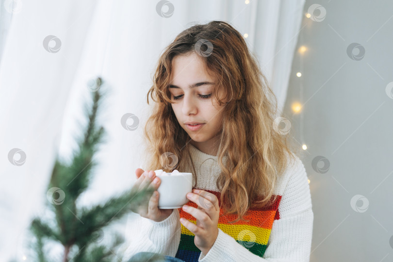 Скачать Красивая девушка-подросток с чашкой какао с зефиром сидит на подоконнике в уютной комнате фотосток Ozero
