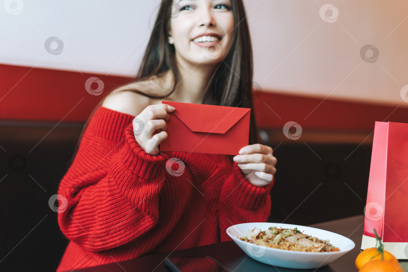 Скачать Красивая молодая азиатка в красной одежде ест азиатскую еду с красным конвертом в ресторане, празднуя китайский Новый год фотосток Ozero