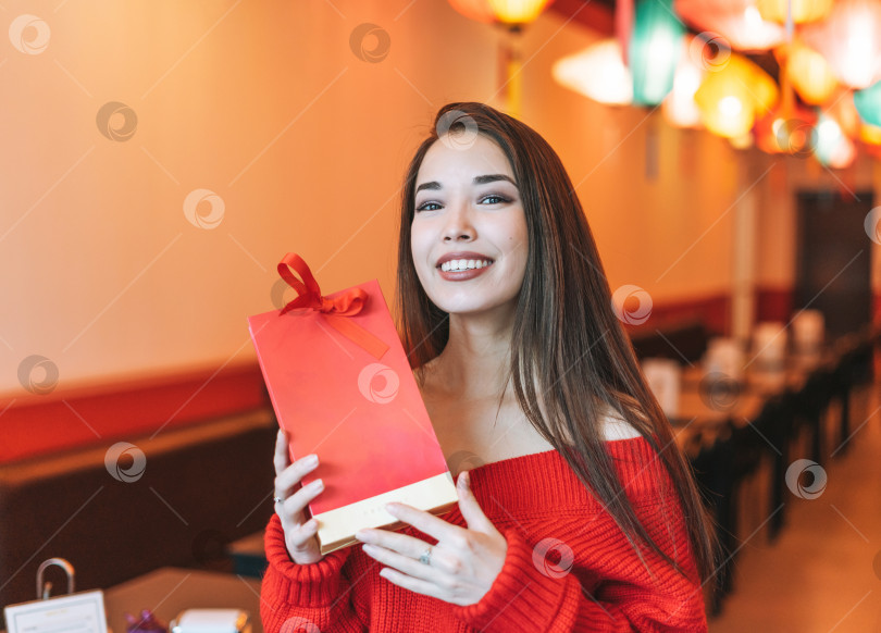 Скачать Красивая молодая азиатка в красной одежде с красным подарочным пакетом в ресторане, празднующая китайский Новый год фотосток Ozero