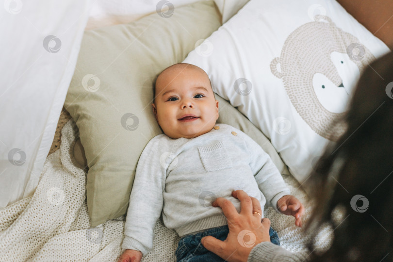 Скачать Милый улыбающийся мальчик бежевого цвета 5-6 месяцев смотрит в камеру вместе с мамой на кровати с вязаным одеялом естественных тонов фотосток Ozero