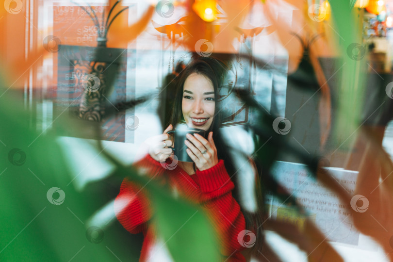 Скачать Красивая улыбающаяся молодая азиатка в красном с чашкой чая в ресторане через окно фотосток Ozero